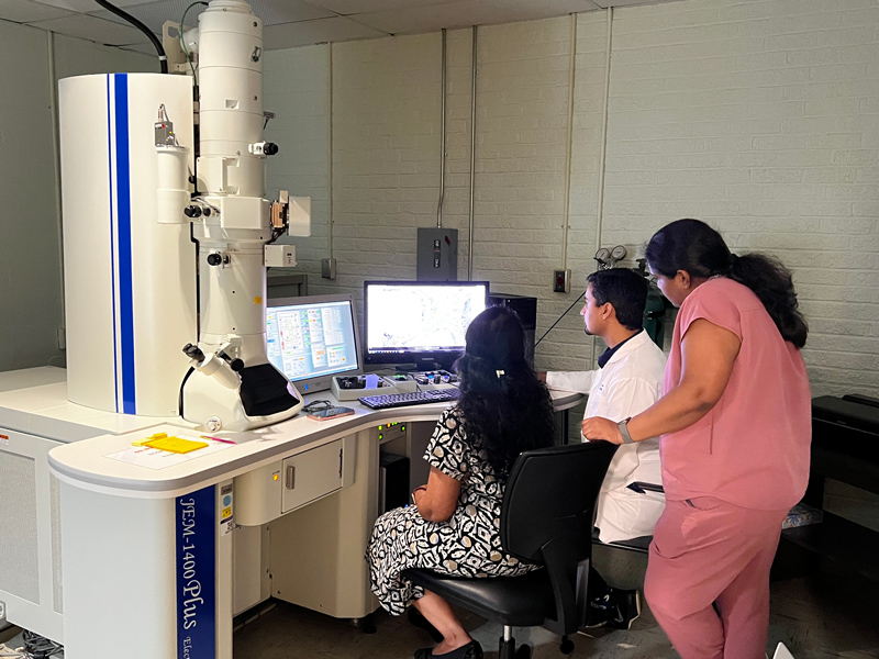 Three people work with a transmission electron microscope, viewing images and data on computer monitors in a laboratory setting.